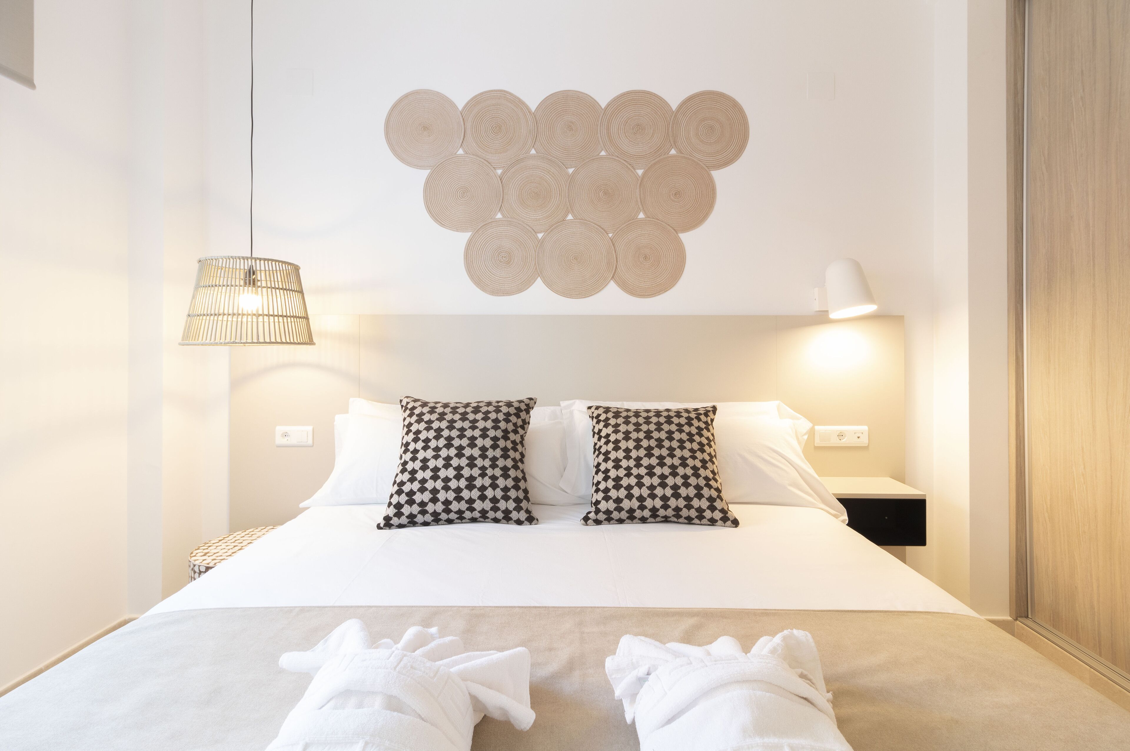 A hotel room with a neatly made bed, two patterned pillows, a wall-mounted circular light fixture, and a bedside lamp.