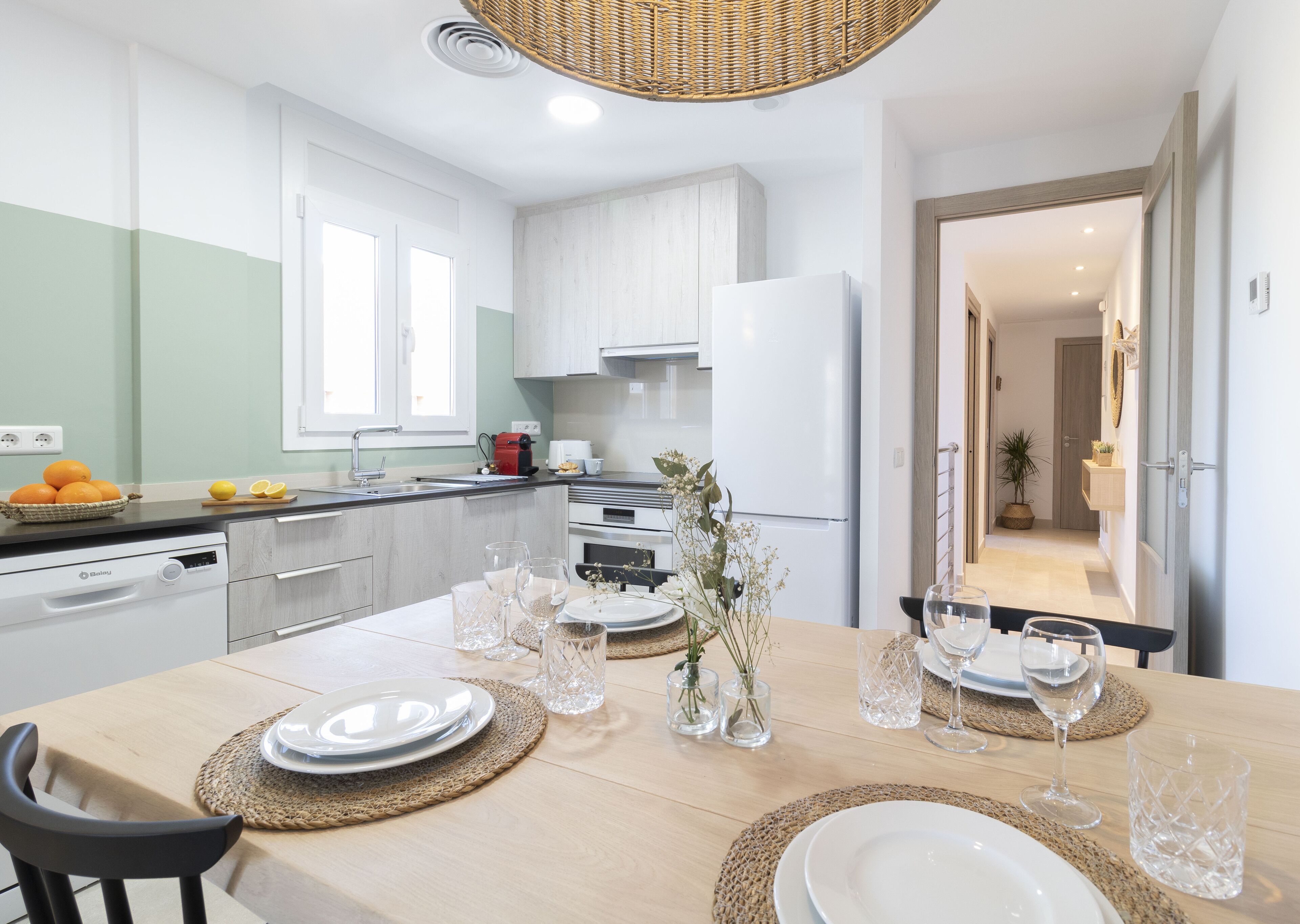 A modern kitchen with white cabinets, a dining table set with plates and glasses, and a wicker ceiling light.