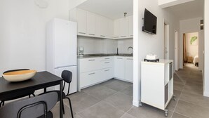 Cocina moderna con armarios blancos, encimera negra y refrigerador blanco. Hay una mesa de comedor con sillas y un tazón encima. Un televisor está montado en la pared.