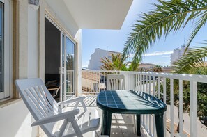 A balcony with white chairs and a table, a sliding glass door, and a view of buildings and palm trees.