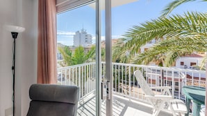 Un balcón con vistas a edificios y palmeras, una silla, una mesa y una lámpara.