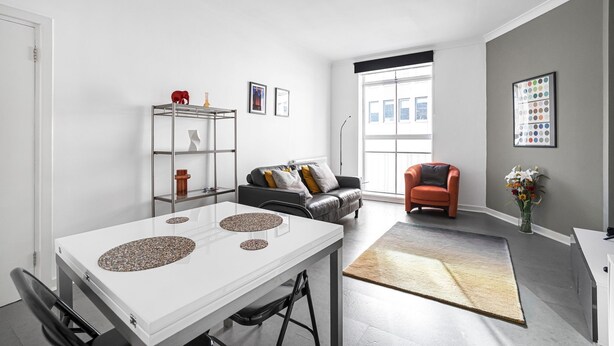 A modern living room with a white dining table, a black leather sofa, and a red armchair. There is a large window providing natural light, a floor lamp, and a vase with flowers on a rug.