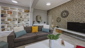 Een moderne woonkamer met een stenen muur, een flatscreen televisie, een grijze bank met decoratieve kussens, en een witte salontafel met een plant.
