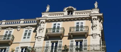 Um edifício com várias andares, varandas ornamentadas, estátuas clássicas e um céu azul claro.