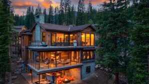 Una cabaña de madera de dos pisos con ventanas grandes y una chimenea, rodeada de un bosque.