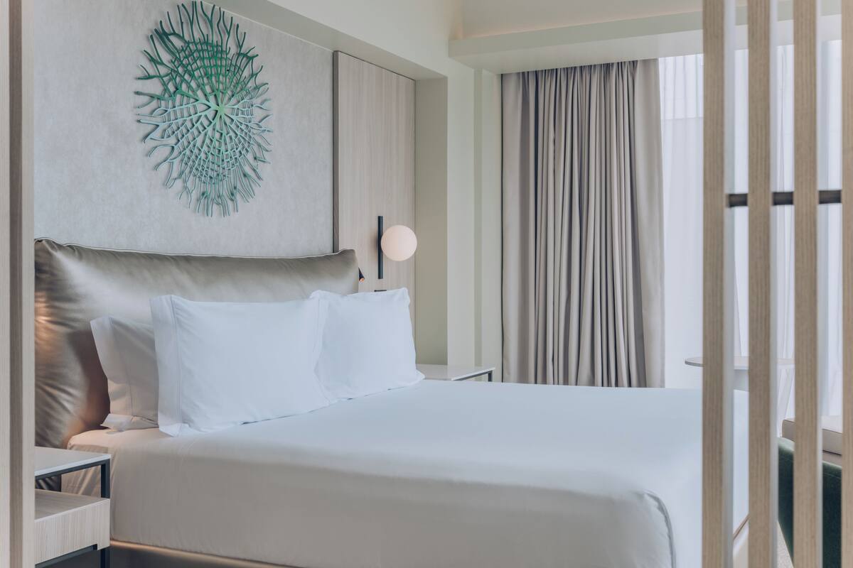 A modern hotel room with a large bed, a decorative wall piece, a bedside table, and a view of the outside through curtains.
