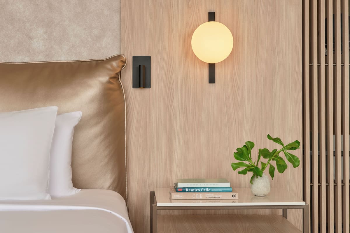 A modern bedroom with a bed, a bedside table with books, a wall-mounted light, and a plant.