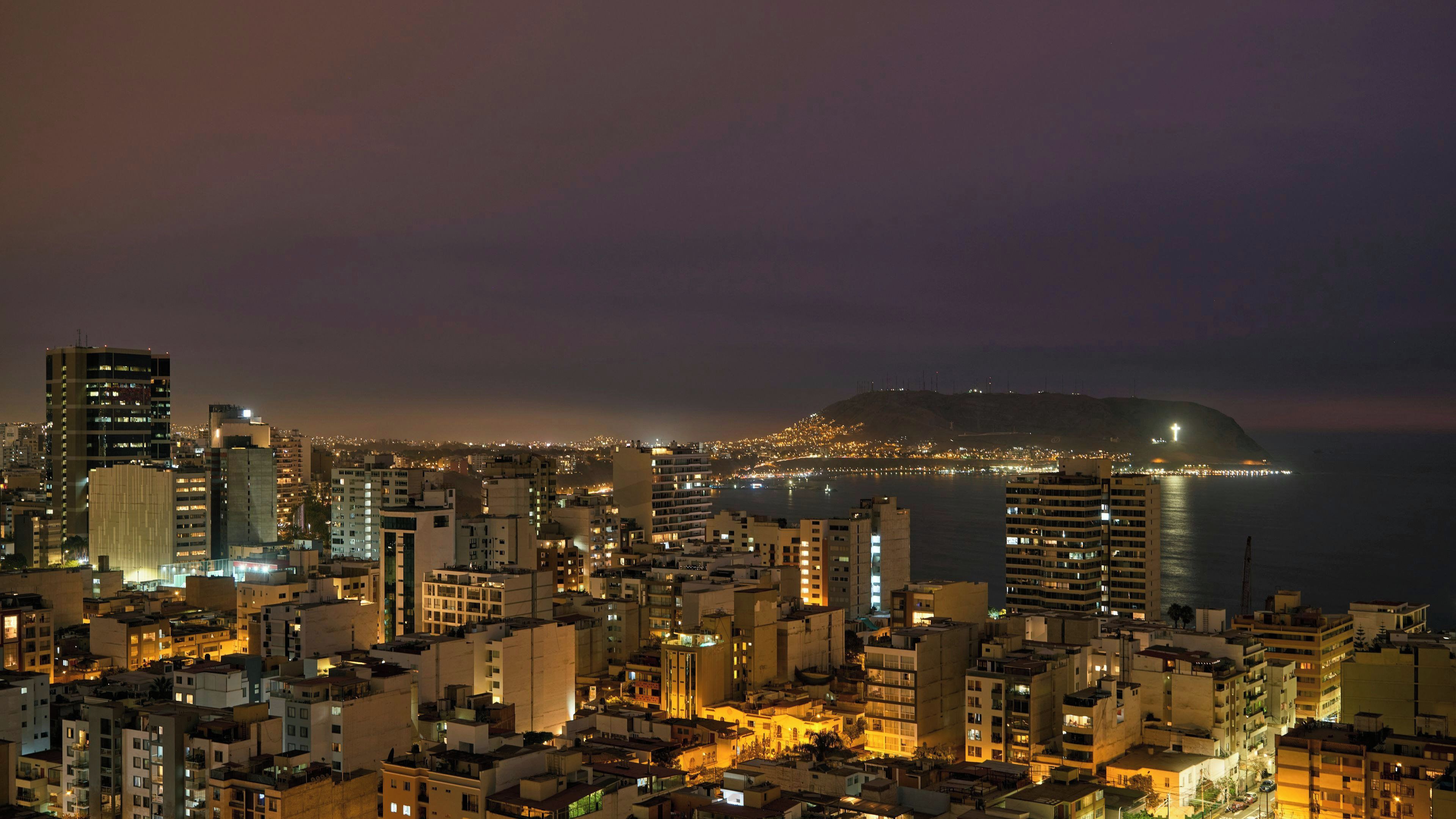A nighttime cityscape with high-rise buildings and a prominent hill with a cross on top.