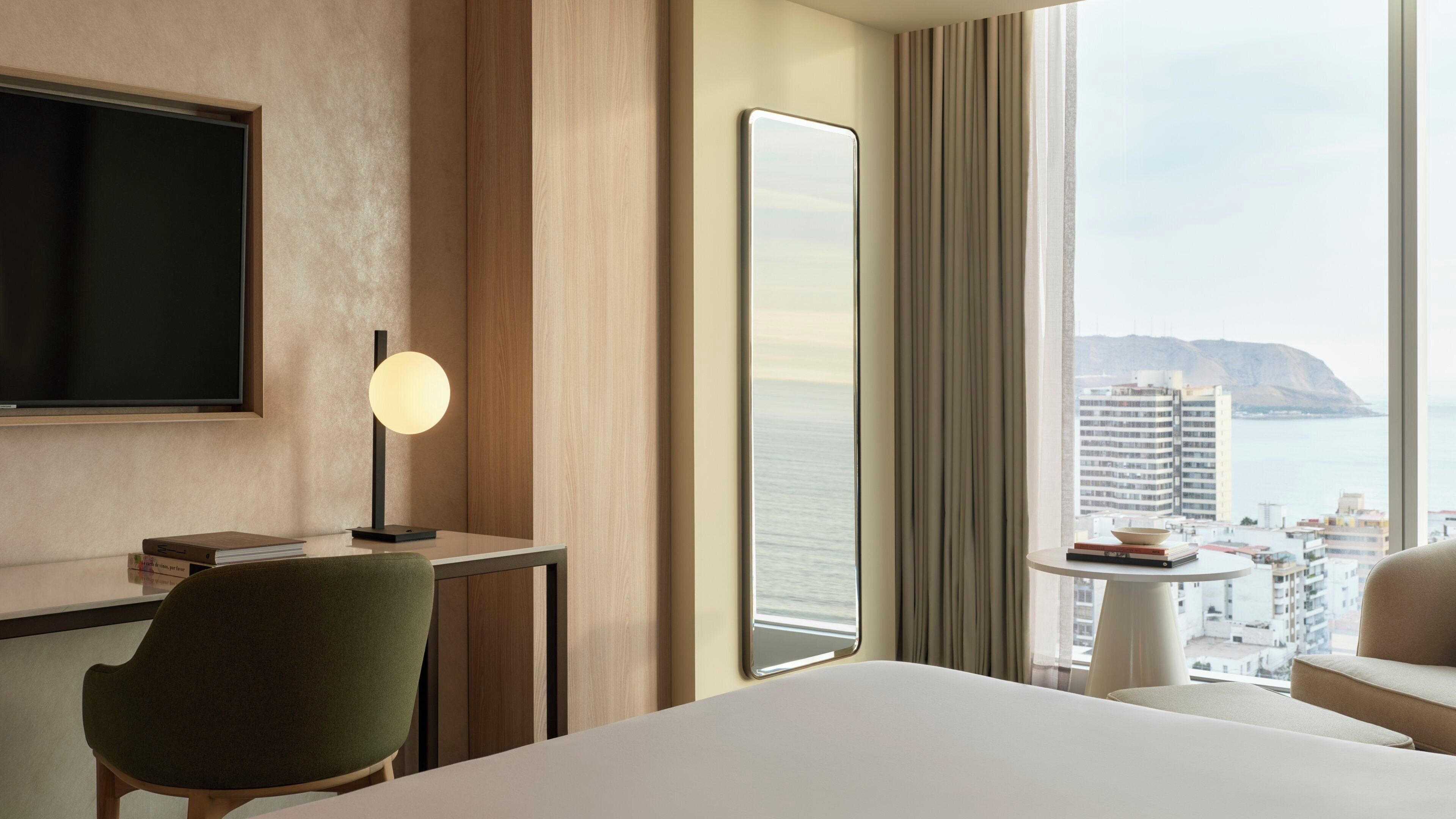 Habitación de hotel con cama, escritorio con lámpara, silla, televisor y vista a la ciudad y al mar.