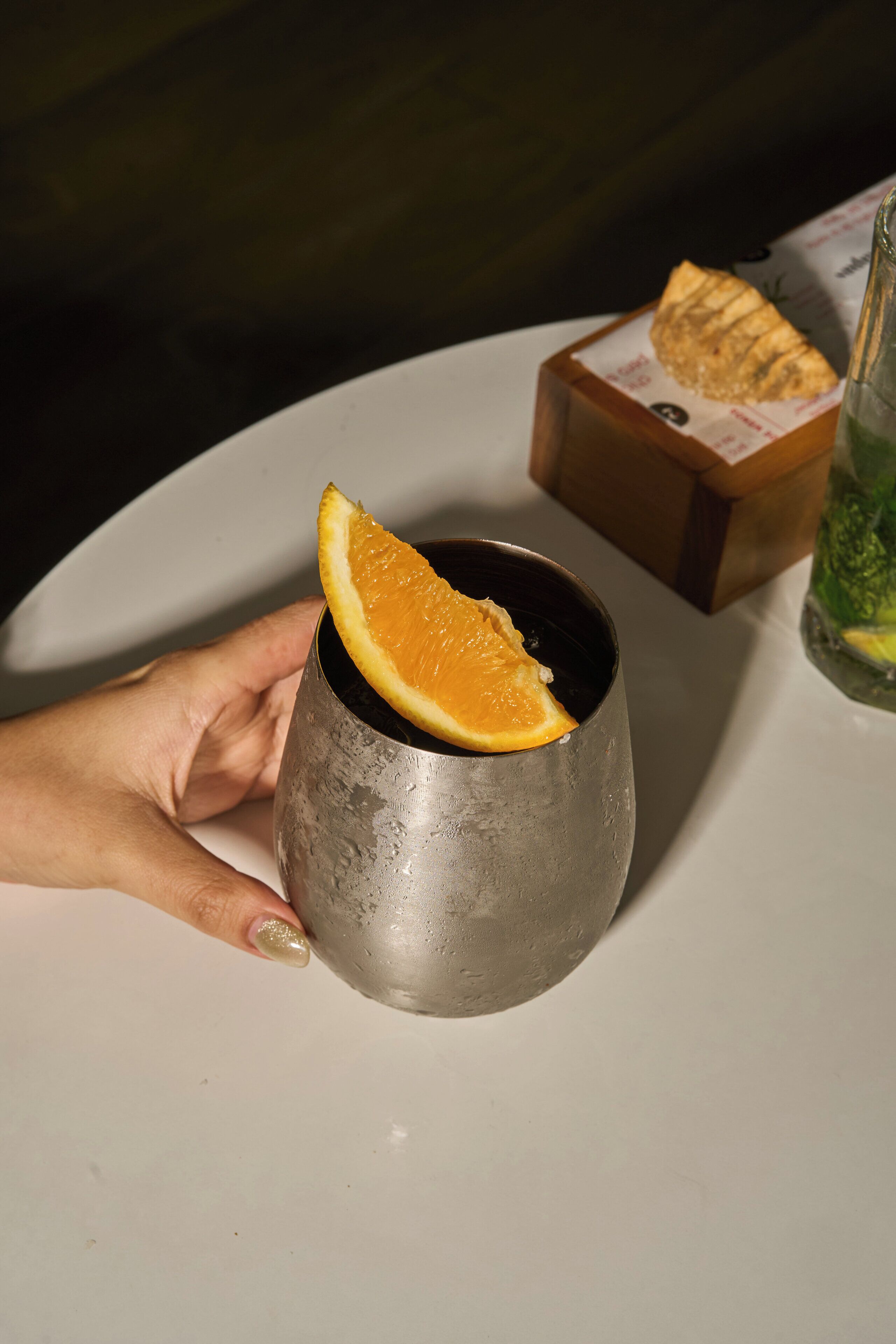 Una mano sosteniendo una taza metálica con una rodaja de naranja.
