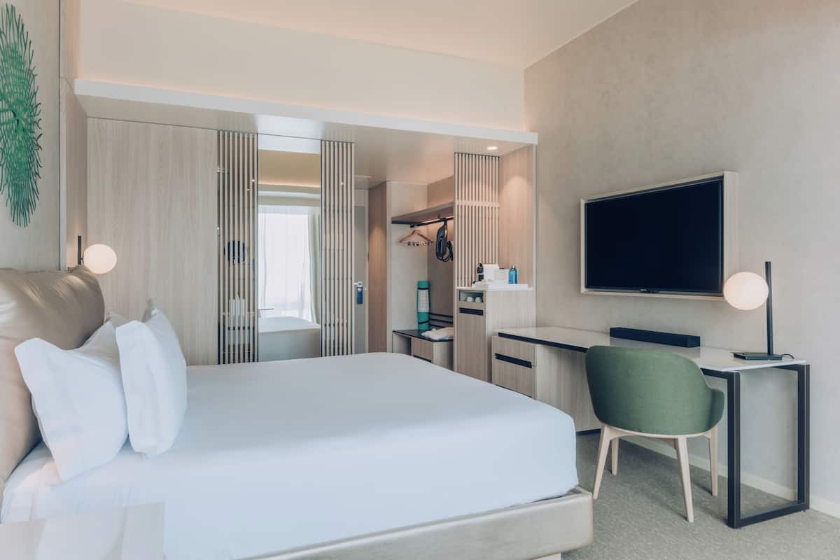 A modern hotel room with a large bed, a desk, a chair, a TV, and a wardrobe.