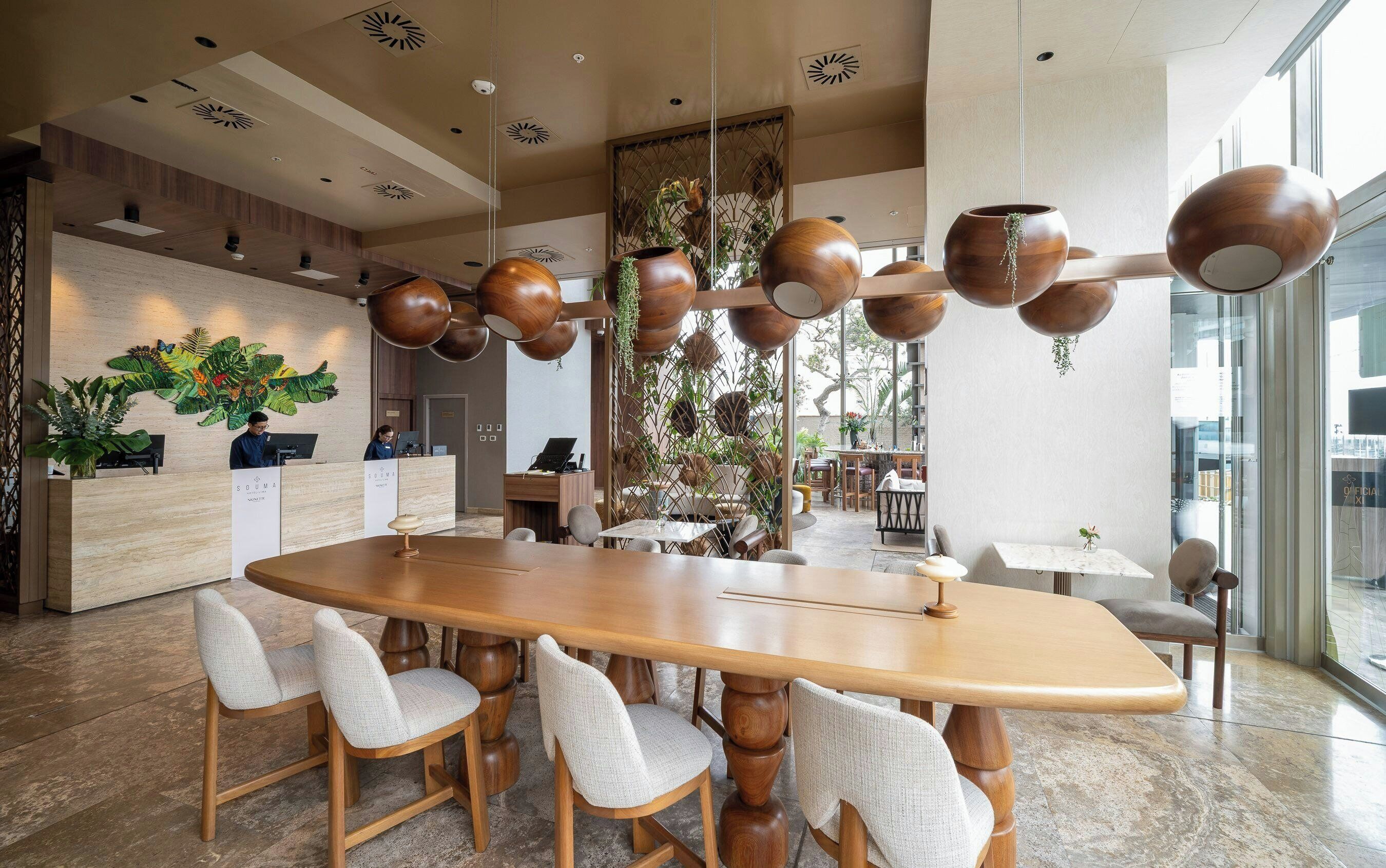 Um lobby de hotel moderno com um balcão de recepção de madeira, uma grande mesa de jantar com cadeiras e um teto decorativo com vasos suspensos.