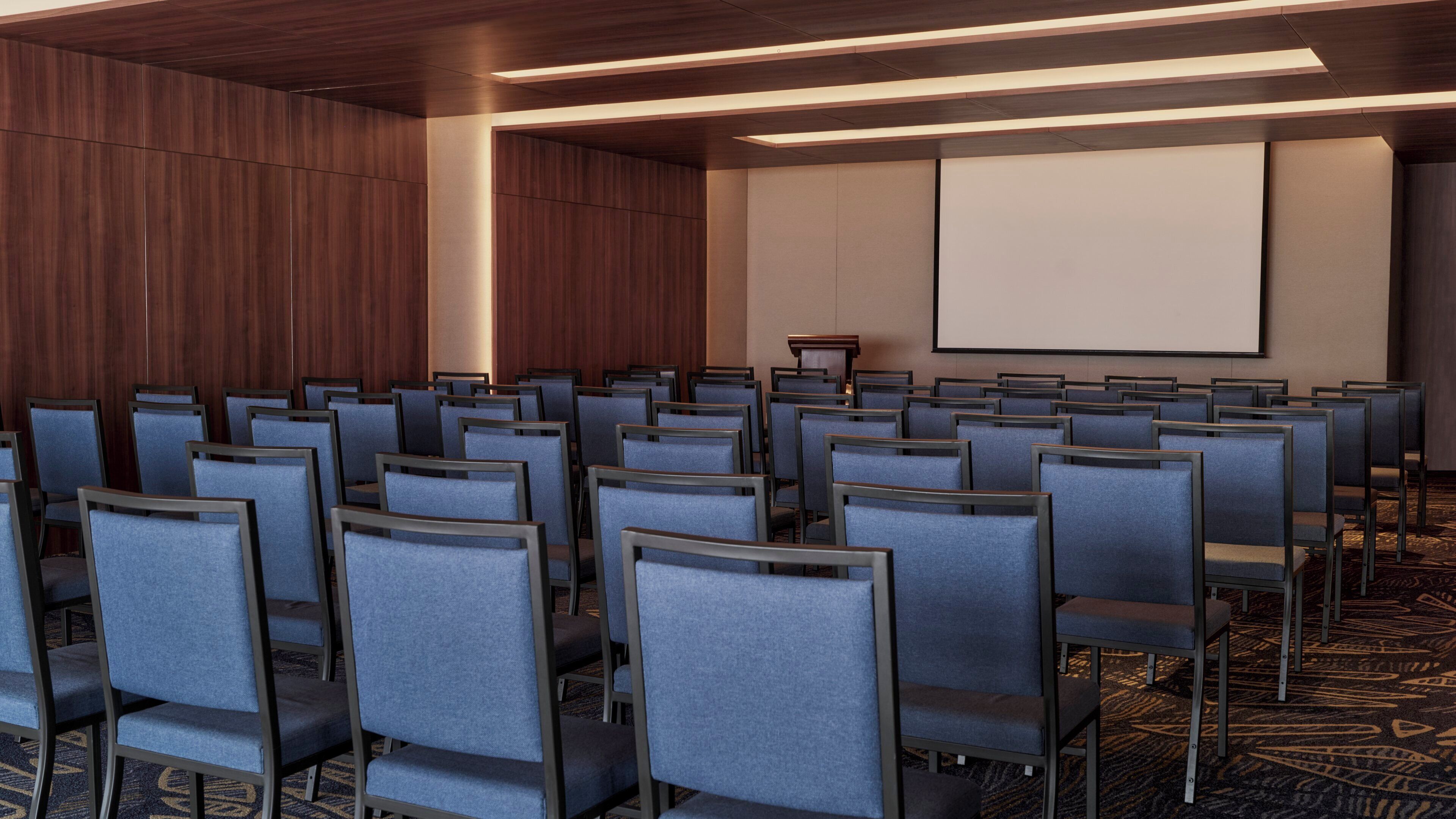 Un salón de conferencias con filas de sillas, un atril y una pantalla de proyección.