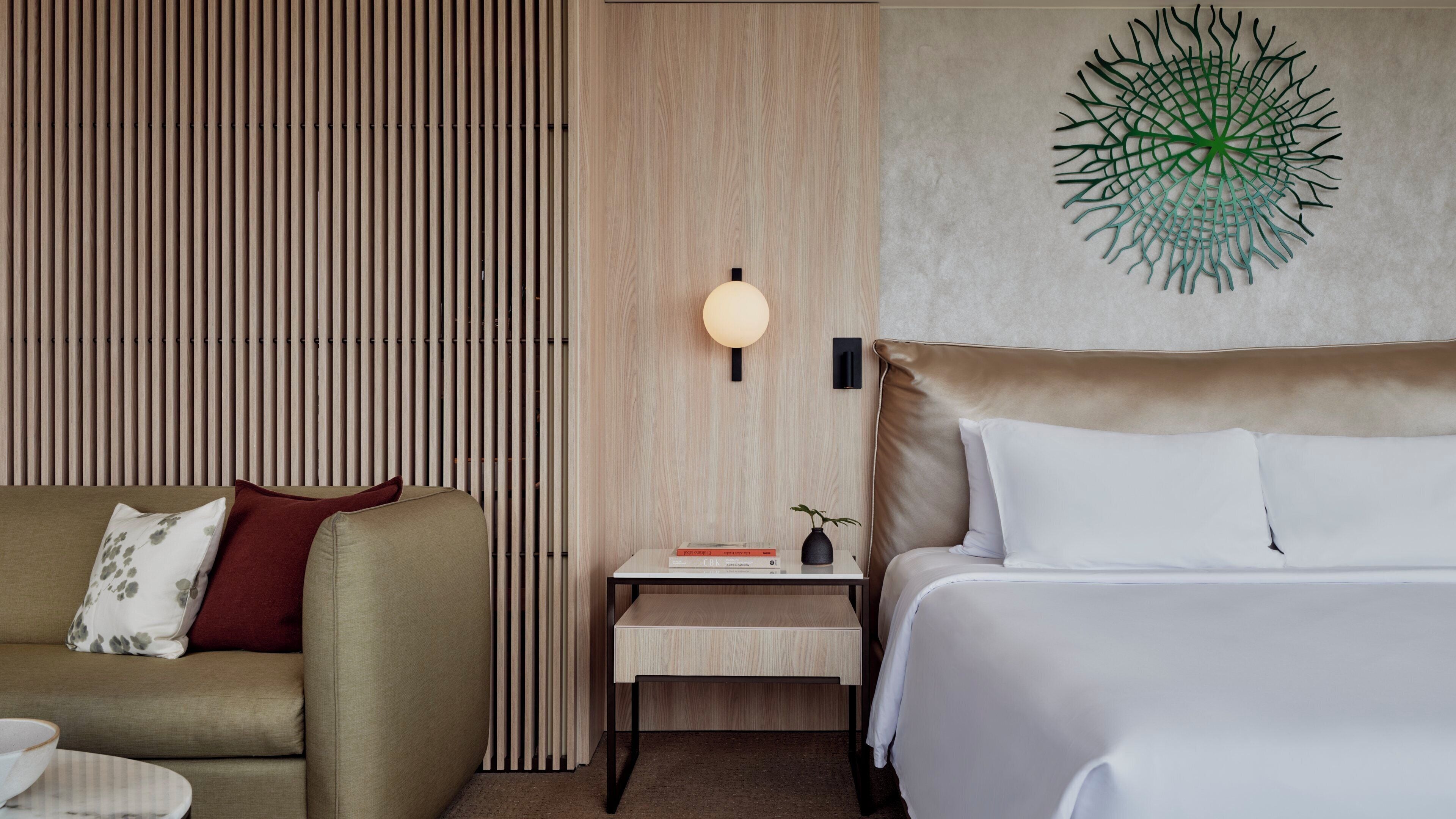 A hotel room with a bed, a sofa, a nightstand with a lamp, and a wall art piece.