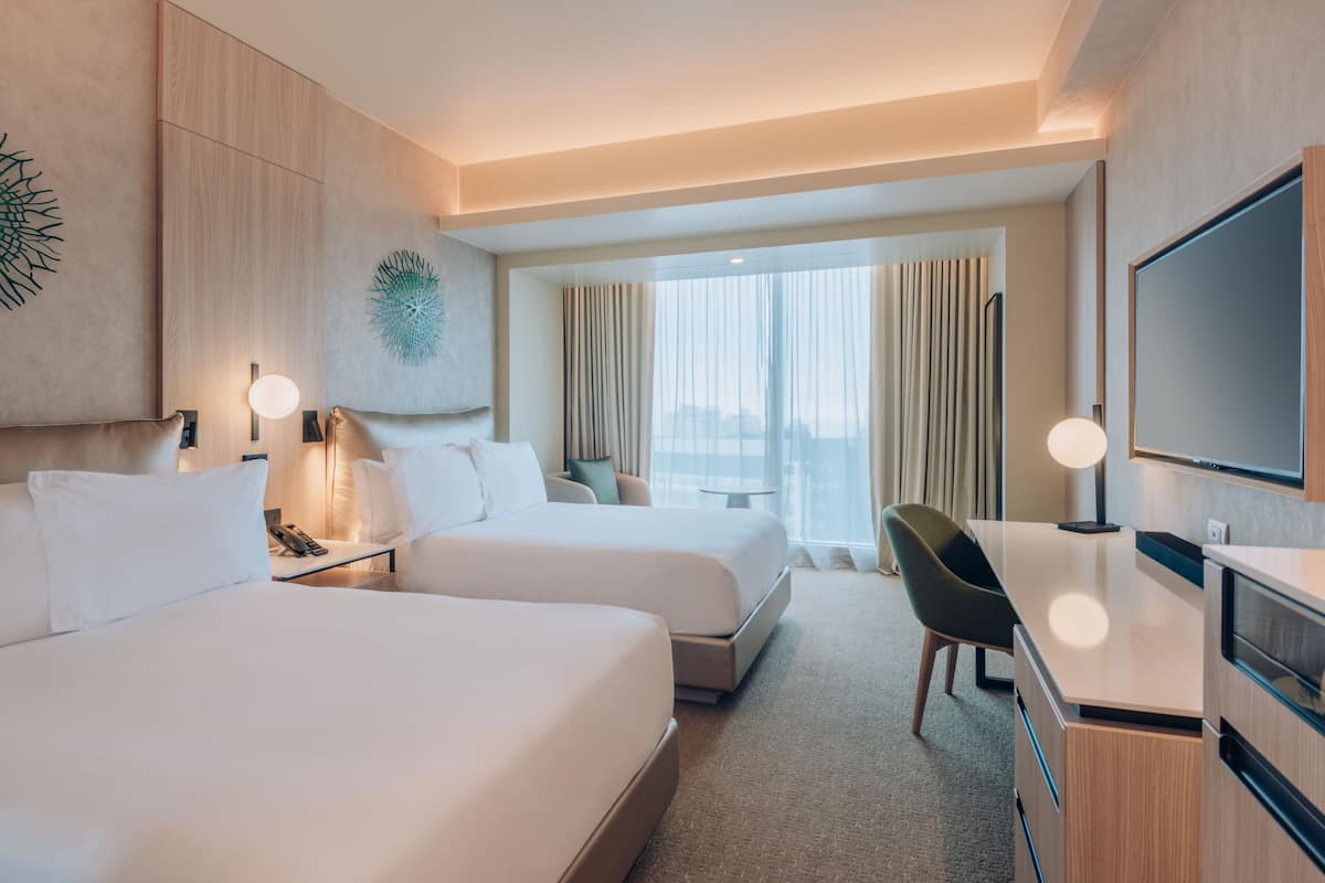 A hotel room with two beds, a desk, a chair, a TV, and a large window with curtains.