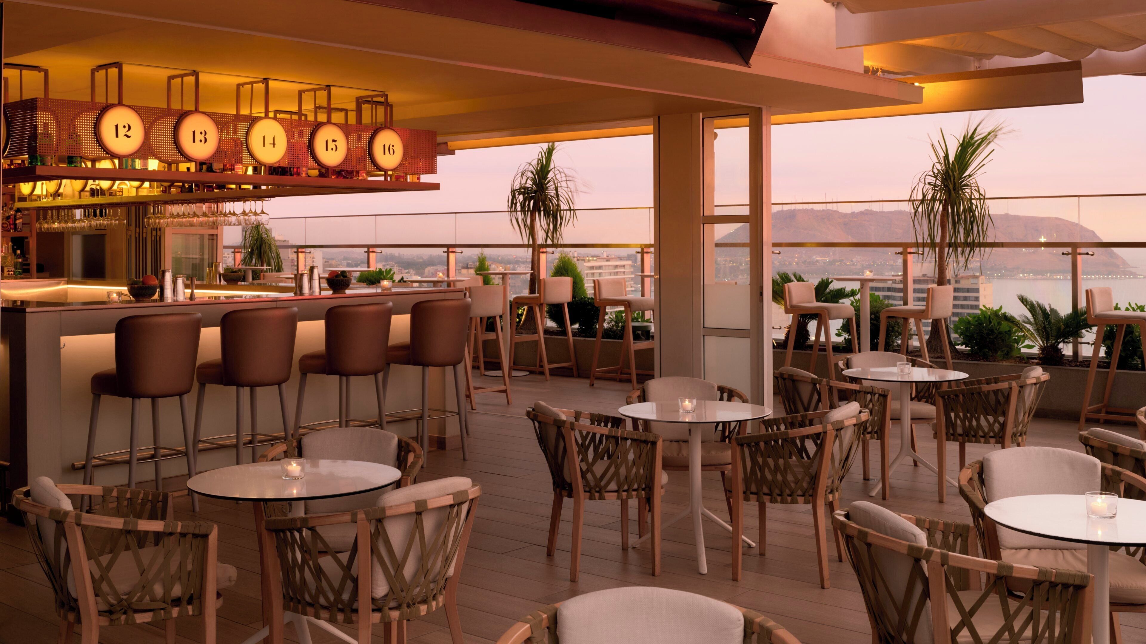 A rooftop bar with multiple seating areas, a bar counter with numbered lights, and a view of the cityscape.