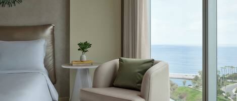 A hotel room with a bed, a small round table with a plant and books, a chair with a pillow, and a large window with a view of the ocean and a bridge.