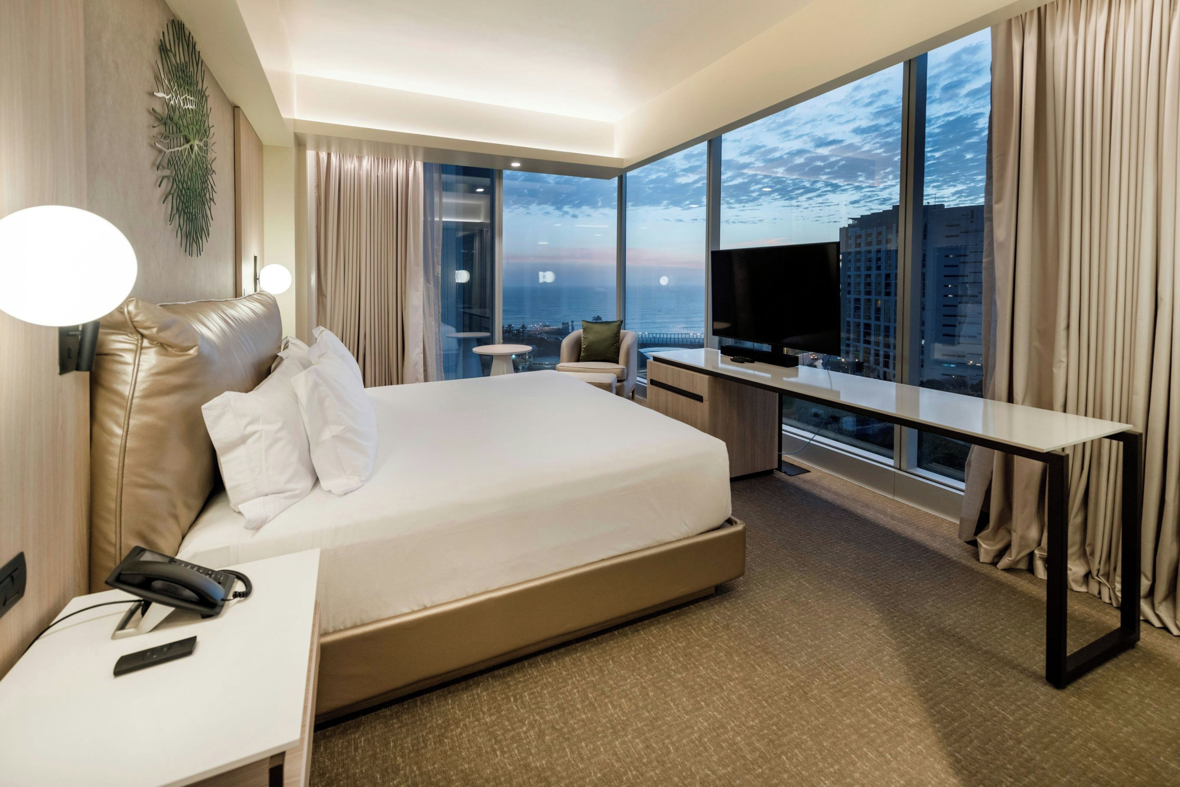 A hotel room with a large bed, a desk, a TV, and a view of the cityscape.