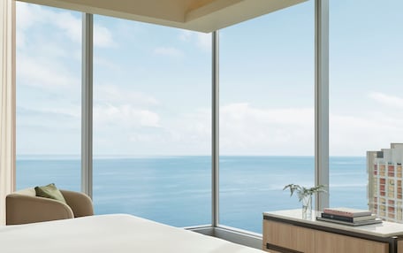 A modern hotel room with a large panoramic window offering a view of the sea and a distant cityscape.