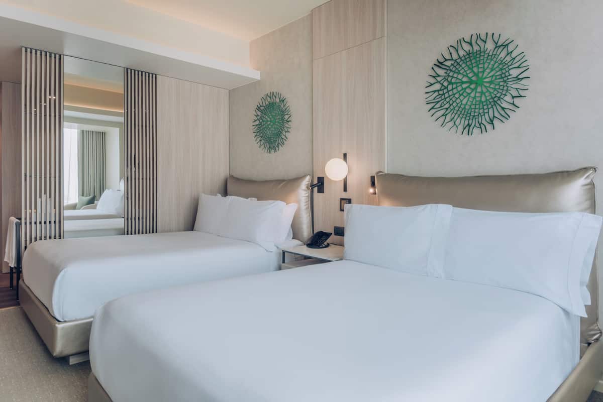 A hotel room with two beds, a mirror, a wall art piece, and a bedside table with a lamp.
