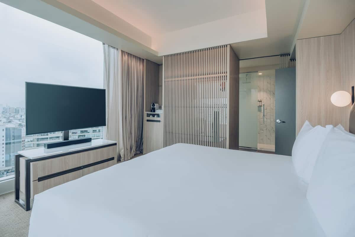 A modern hotel room with a large bed, a flat-screen TV on a wooden console, a walk-in closet, and a city view.