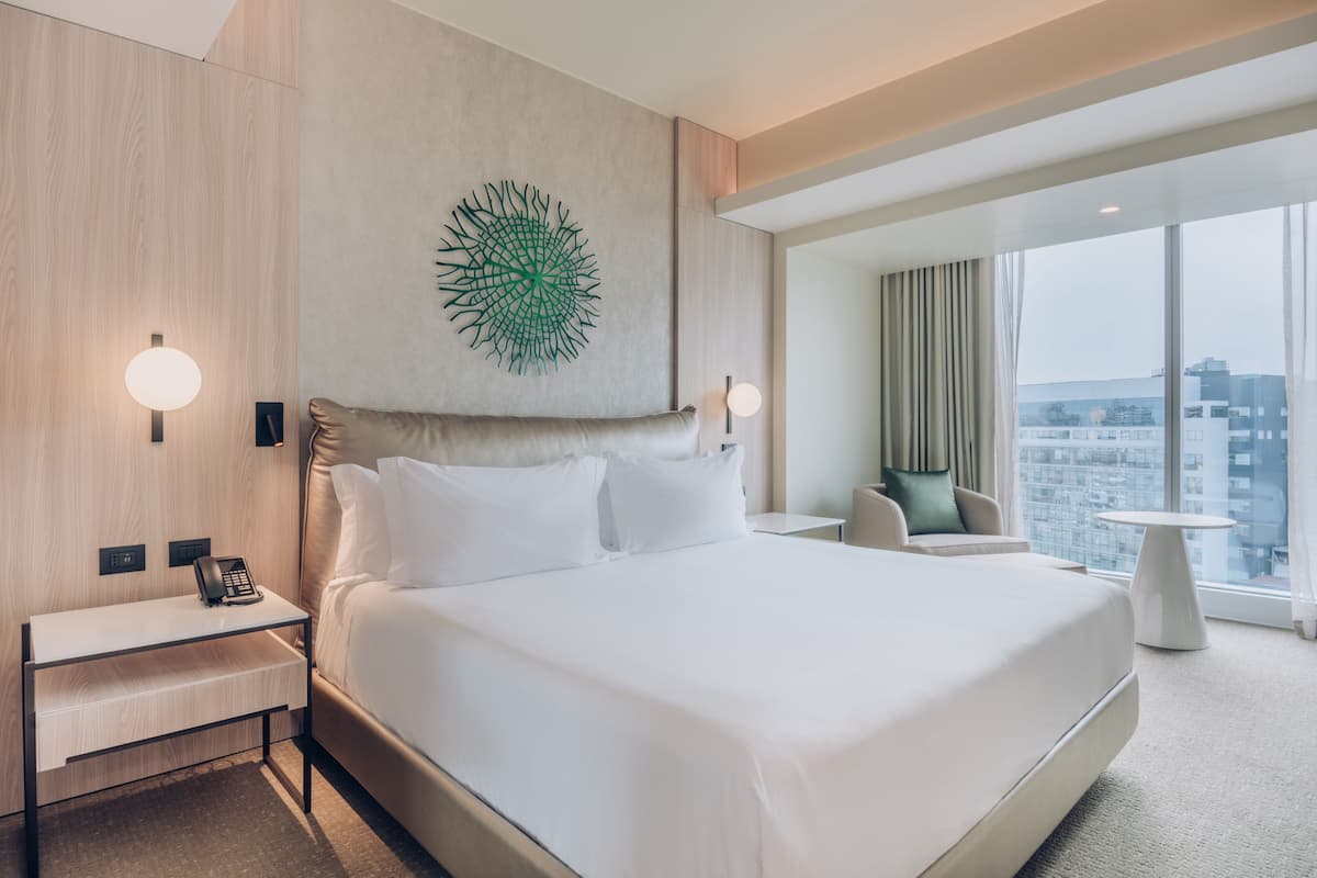 A modern hotel room with a large bed, a desk with a phone, a chair, a small table, and a large window with a city view.