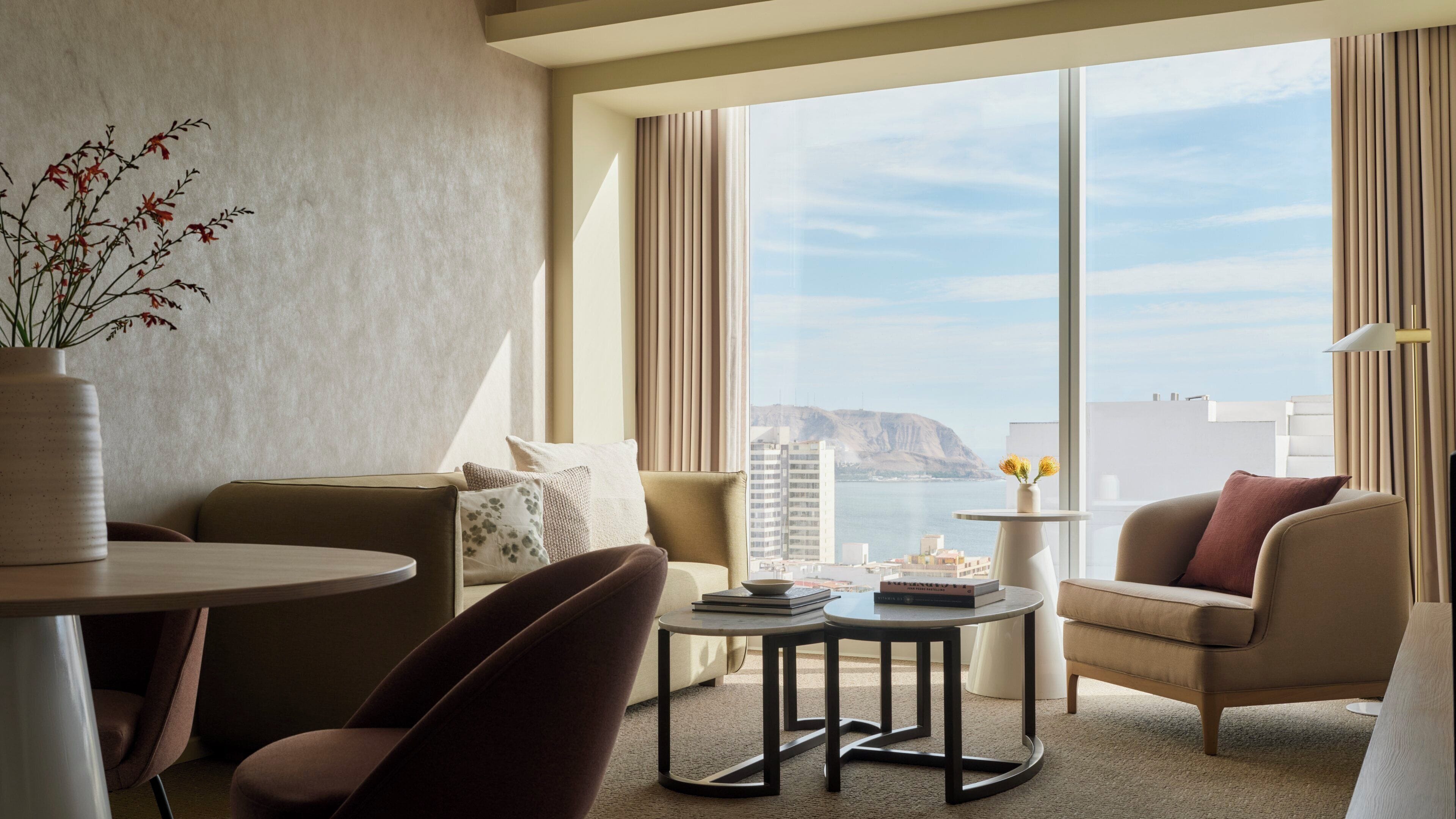 Una habitación de hotel moderna con un ventanal grande, un sofá, una silla, una mesita y un florero con flores.