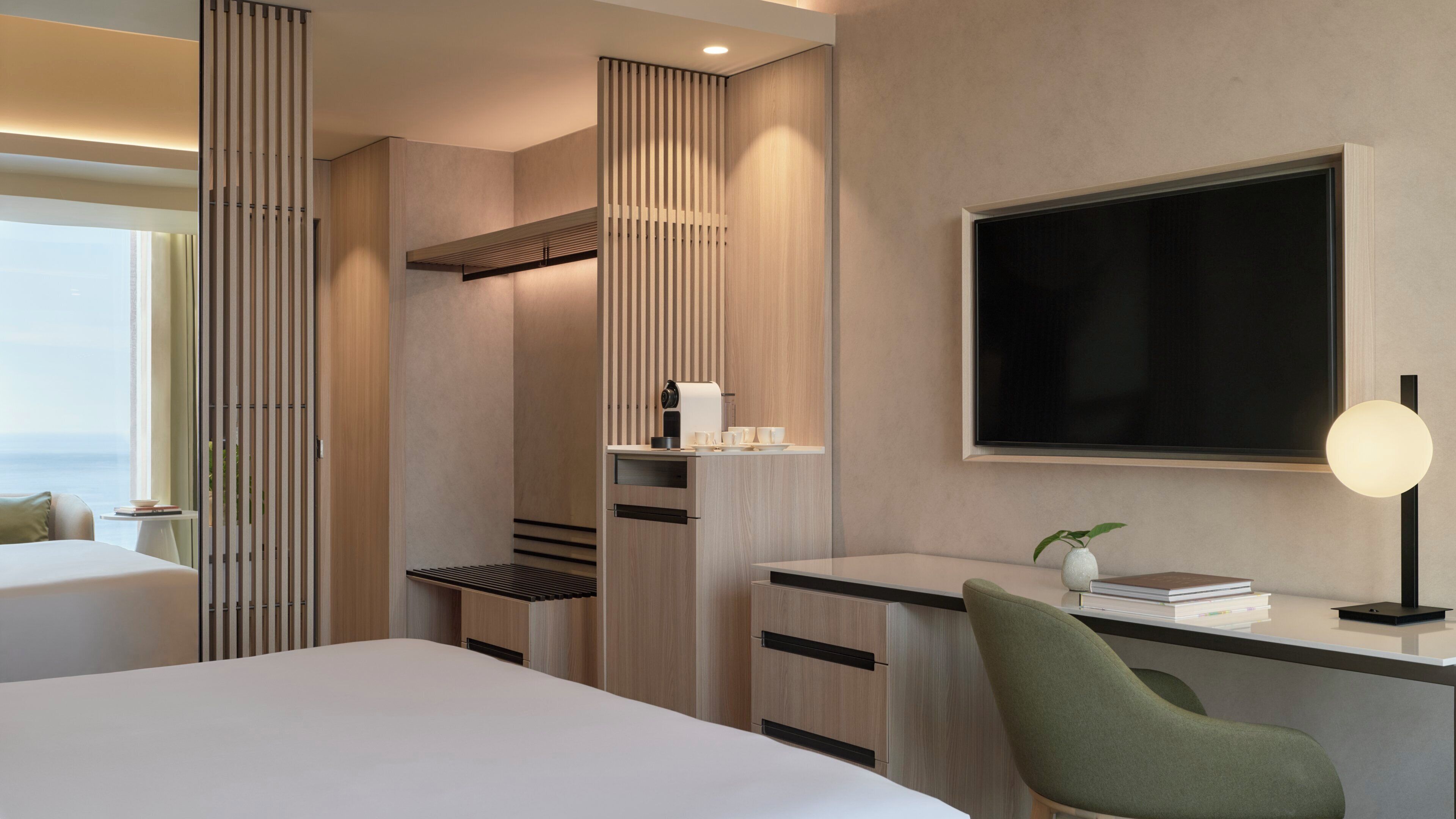 Una habitación de hotel moderna con cama, escritorio, silla, televisor de pantalla plana y mesita de noche con lámpara.