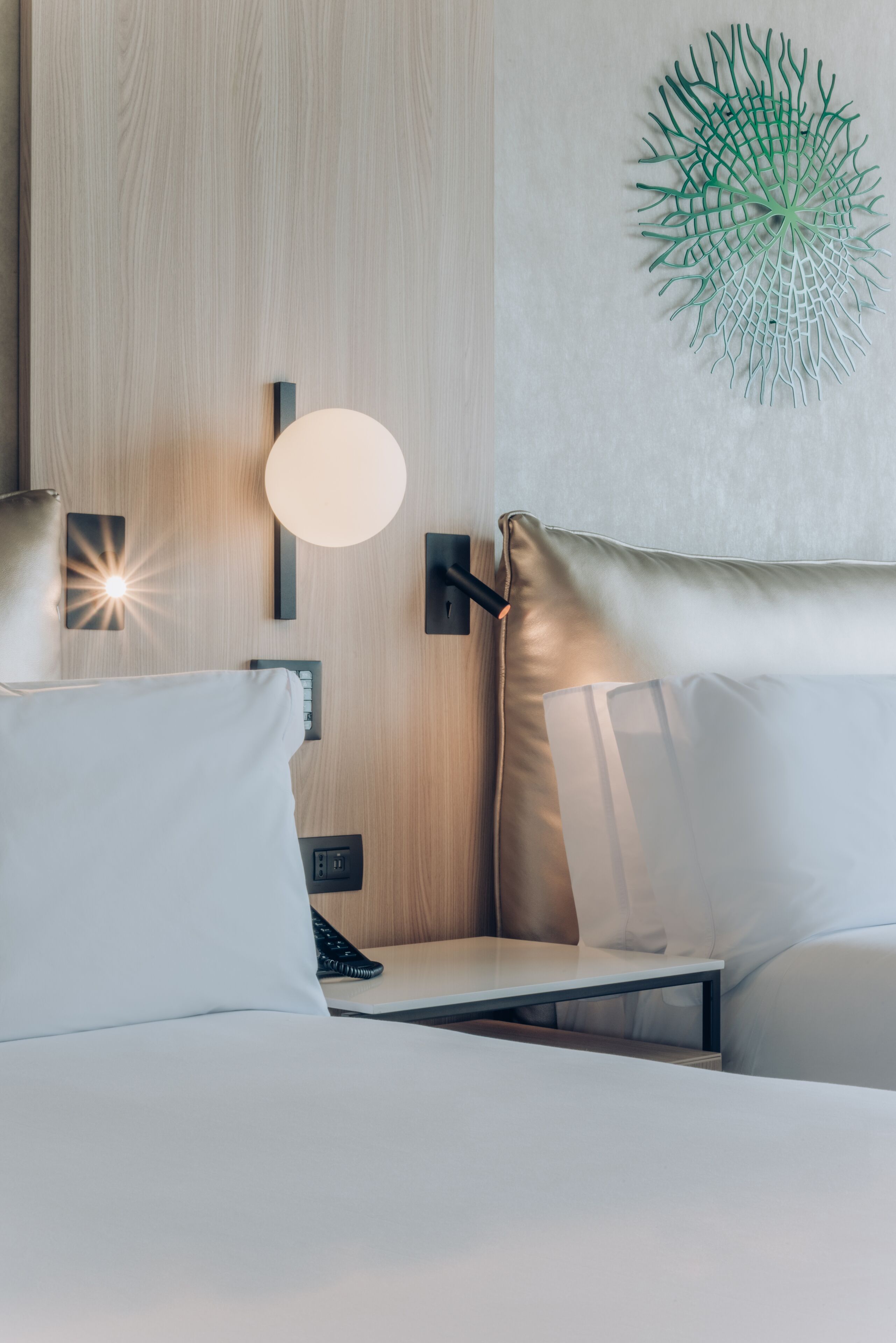 A modern hotel room with a wooden headboard, a round wall light, a decorative wall piece, and a bed with white and beige bedding.