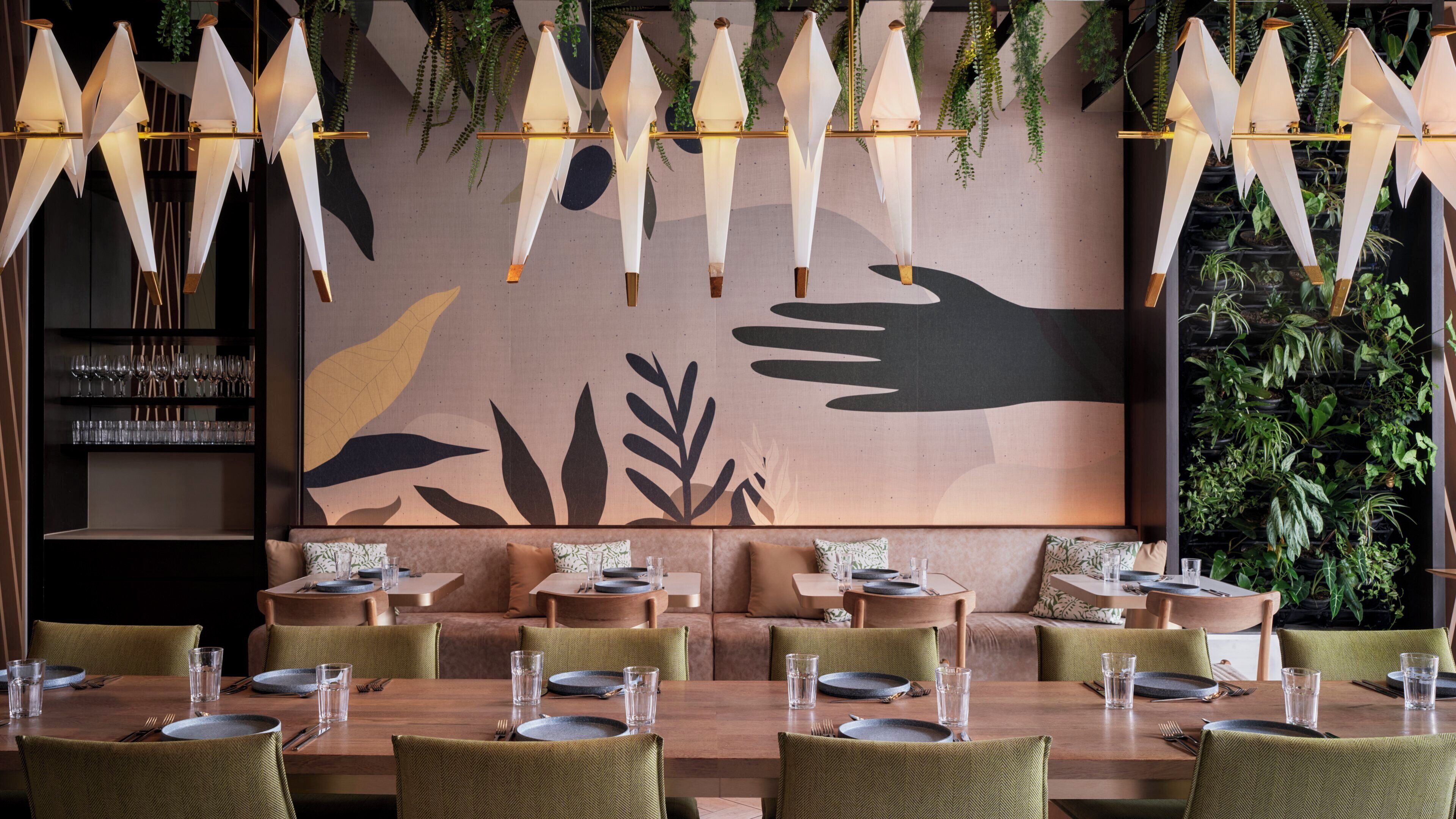 A restaurant interior with a long table set for dining, a wall with a large abstract painting, and hanging plants.