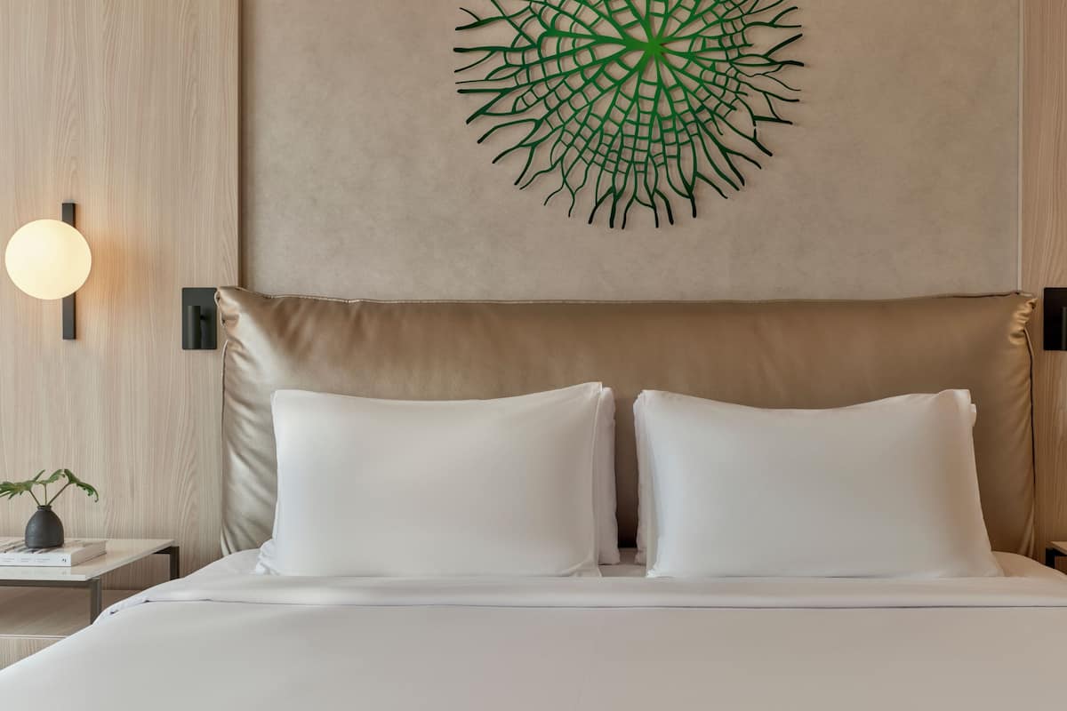 A modern hotel room with a large bed, two white pillows, a wall-mounted green abstract art piece, and two wall sconces.