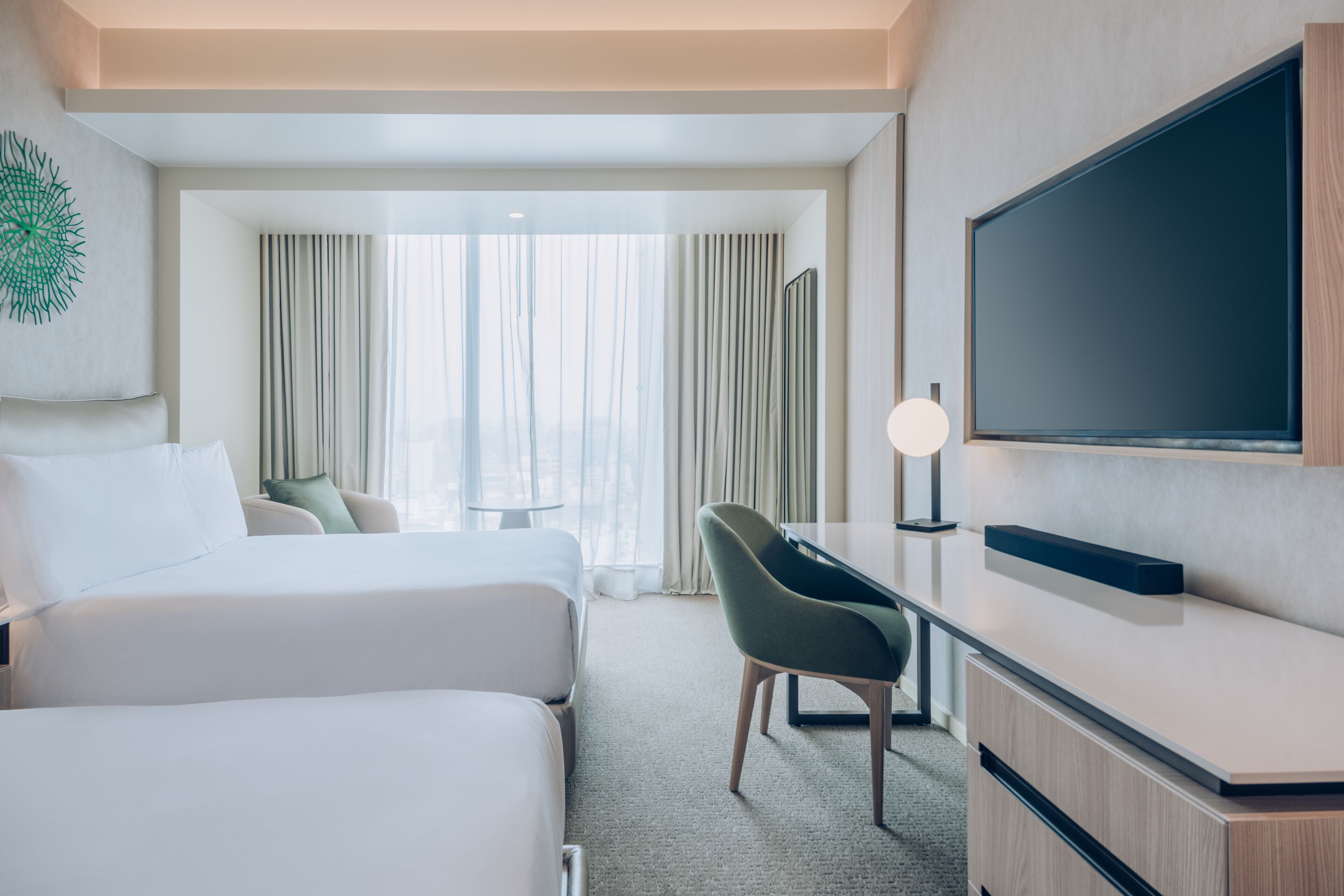 A hotel room with two beds, a desk, a TV, and a view of the city through large windows.