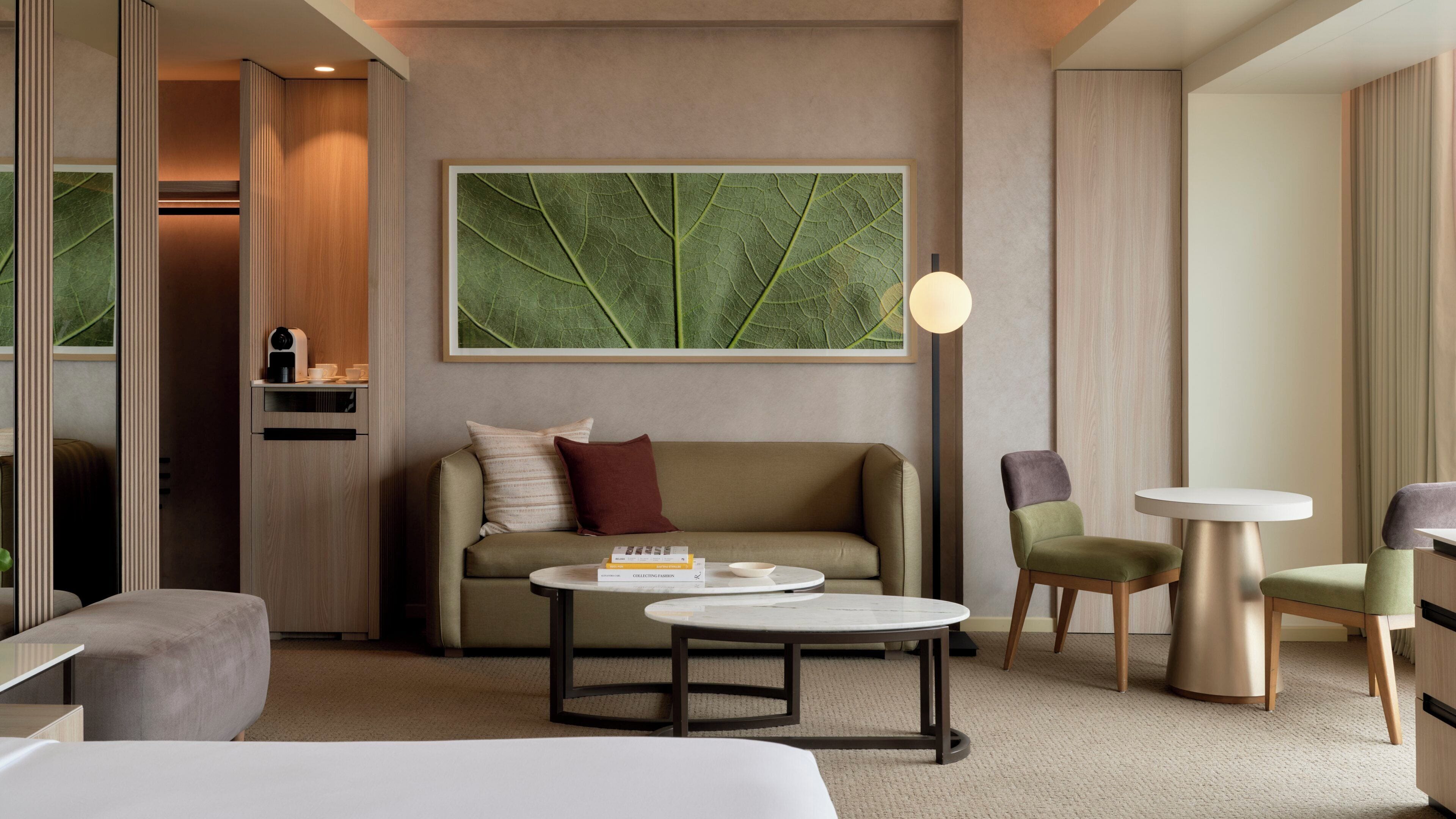 Una habitación de hotel moderna con sofá, dos sillones, una mesita de café, una mesita auxiliar, una lámpara y una obra de arte grande con estampado de hojas en la pared.
