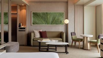 A modern hotel room with a sofa, two armchairs, a coffee table, a side table, a lamp, and a large leaf-patterned artwork on the wall.