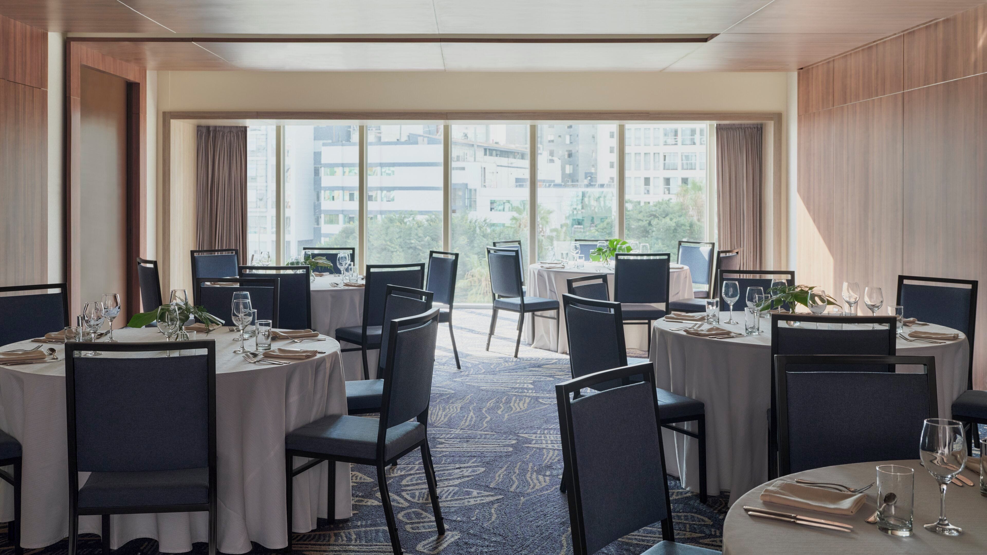 A hotel dining area with multiple round tables, chairs, and a view of buildings through large windows.