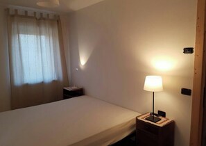 Une chambre d’hôtel comprenant un lit, une table de chevet avec une lampe, une fenêtre avec des rideaux et un luminaire fixé au mur.