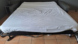 Valkoisella patjalla ja mustalla rungolla varustettu sänky, pieni yöpöytä, jossa on lamppu, ja harmaa seinä.