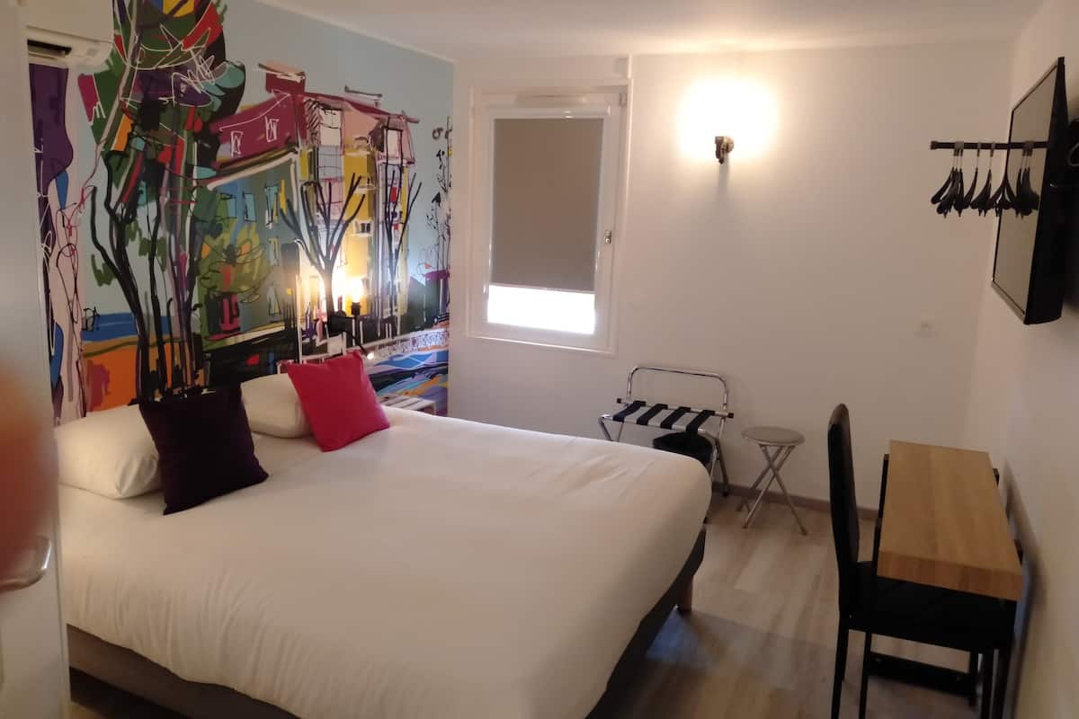 A bedroom with a large bed, a small table, a chair, a wall-mounted rack, and a window.