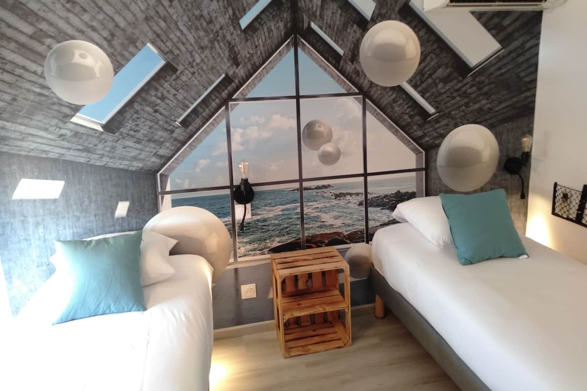 A bedroom with a large window, a bed, a wooden stool, and a wall-mounted lamp.