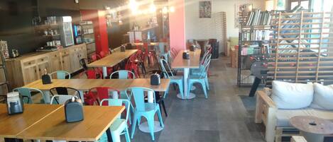 A restaurant with wooden tables and chairs, a counter with kitchen equipment, and a bookshelf with books.