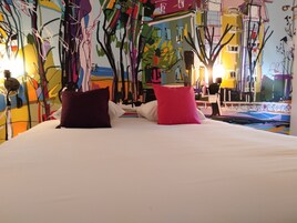 Una cama con dos almohadas, un cuadro abstracto y colorido en la pared, y una ventana con vistas a edificios.
