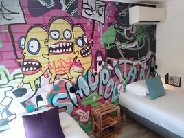 Una habitación con una pared decorada con grafitis coloridos, una cama con colchón blanco y almohadas azul turquesa, un taburete de madera pequeño y un equipo de aire acondicionado instalado en la pared.
