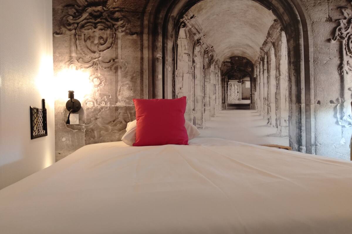 A room with a bed, a red pillow, and a wall with a stone archway leading to another room.