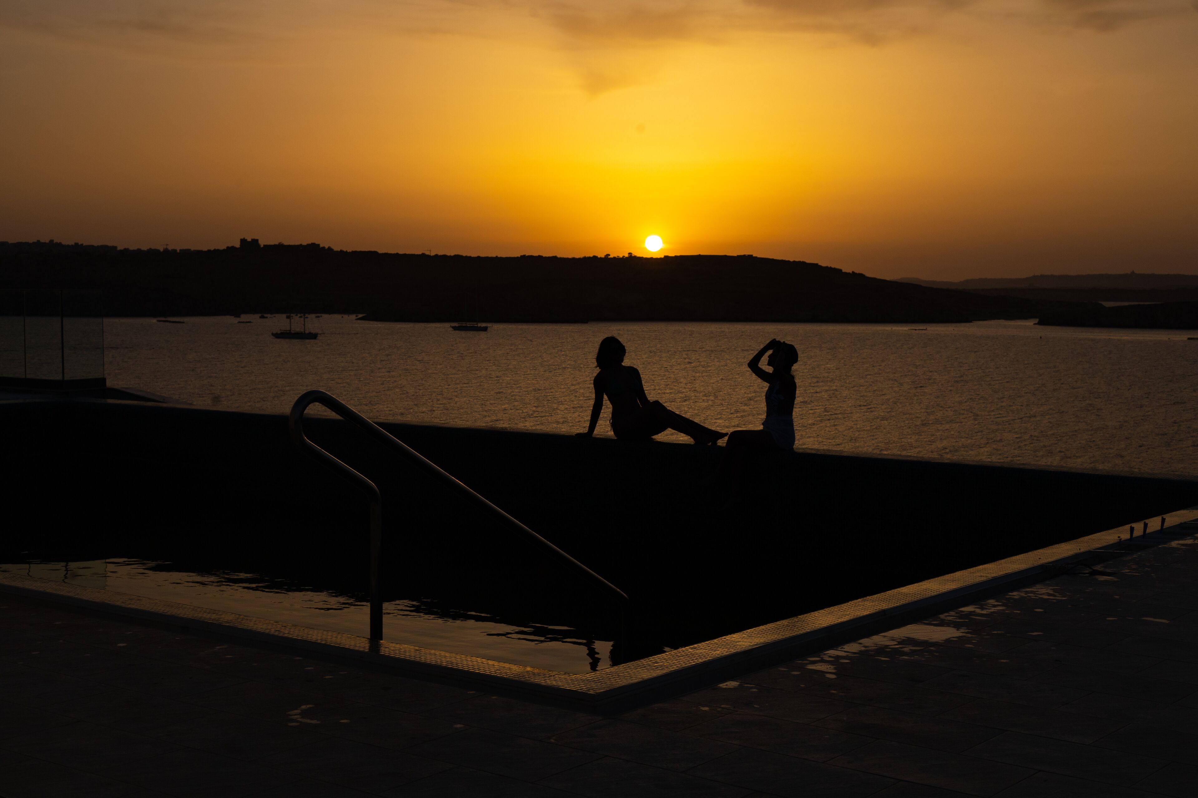 A silhouette of two people at a swimming pool during sunset.