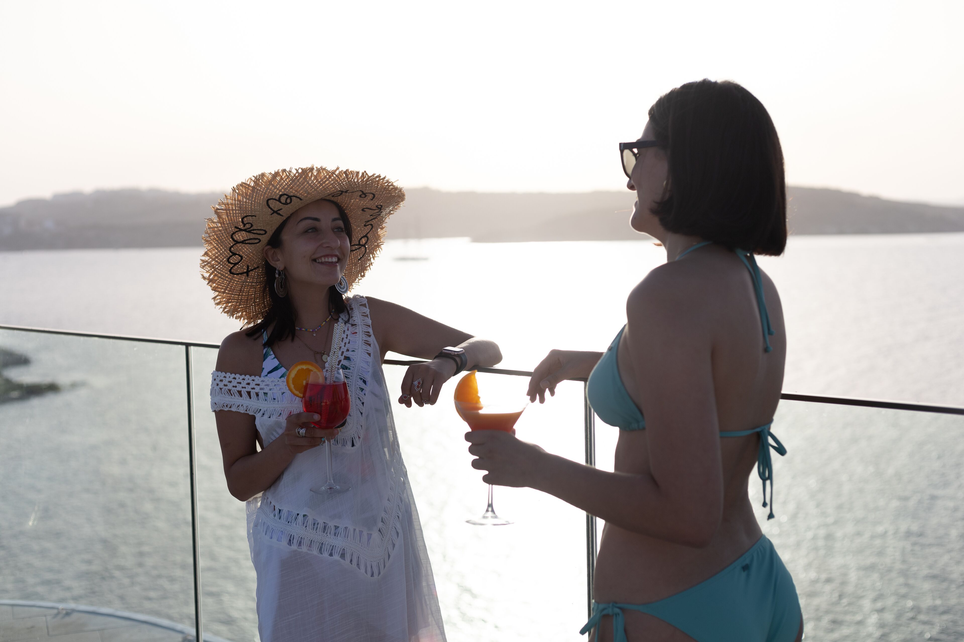 Dos personas en un balcón con vistas a un cuerpo de agua, sosteniendo bebidas.