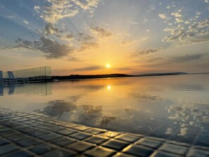 Rolig solnedgang over et stille vandområde med pool og liggestole til venstre.
