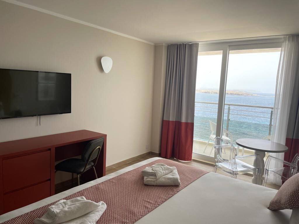 A hotel room with a bed, a desk, a chair, a TV, a balcony with a table and chairs, and a view of the sea.