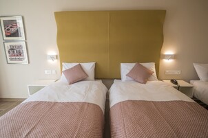 Habitación de hotel con dos camas, cada una con ropa blanca y detalles rojos estampados. Hay luminarias fijadas a la pared a ambos lados de las camas.