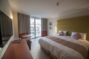 Habitación de hotel con una cama grande, un escritorio con silla, una mesita y vistas al balcón exterior.