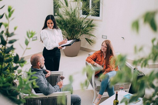 Une scène intérieure moderne : une personne est debout et tient un classeur, une autre est assise sur un canapé avec une boisson, et une troisième personne est assise sur une chaise.